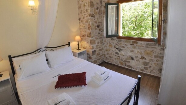 Une chambre avec un mur en pierre, un lit recouvert de draps blancs, un plaid rouge, une table de chevet avec une lampe et une fenêtre donnant sur un paysage verdoyant.