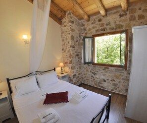 Une chambre avec un mur en pierre, un lit recouvert de draps blancs, un plaid rouge, une table de chevet avec une lampe et une fenêtre donnant sur un paysage verdoyant.