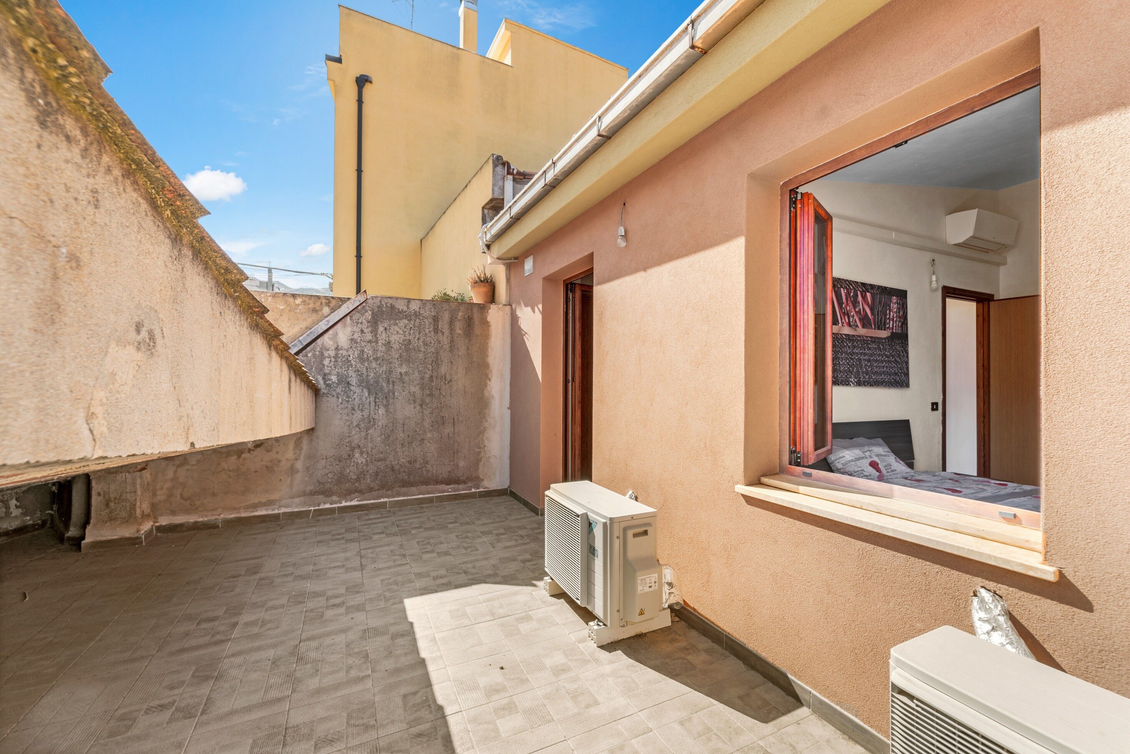 Un pequeño patio estrecho con suelo de baldosas, una ventana con vistas y un equipo de aire acondicionado.