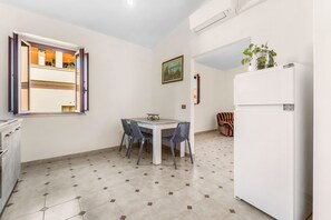 A kitchen with a dining table, chairs, a refrigerator, a window with open shutters, and a painting on the wall.