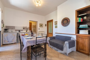 Een eetgedeelte met een tafel voor vier personen, een bank, een boekenkast en een keuken op de achtergrond.
