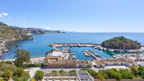 Una marina costiera con imbarcazioni ormeggiate, una piccola isola e edifici dai tetti in terracotta.