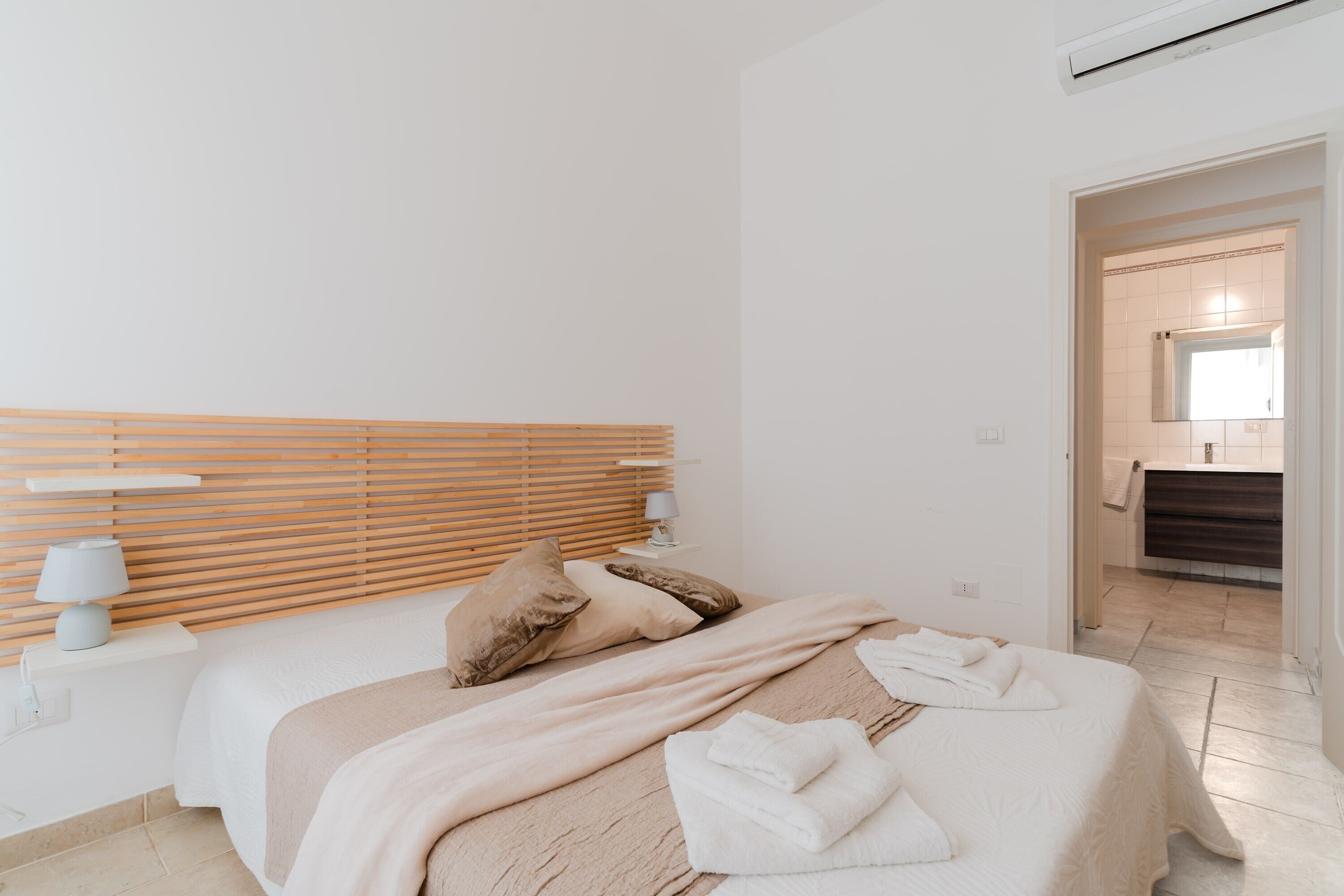 A bedroom with a wooden headboard, a bedside table with a lamp, a bed with a beige blanket, and a bathroom visible through an open door.