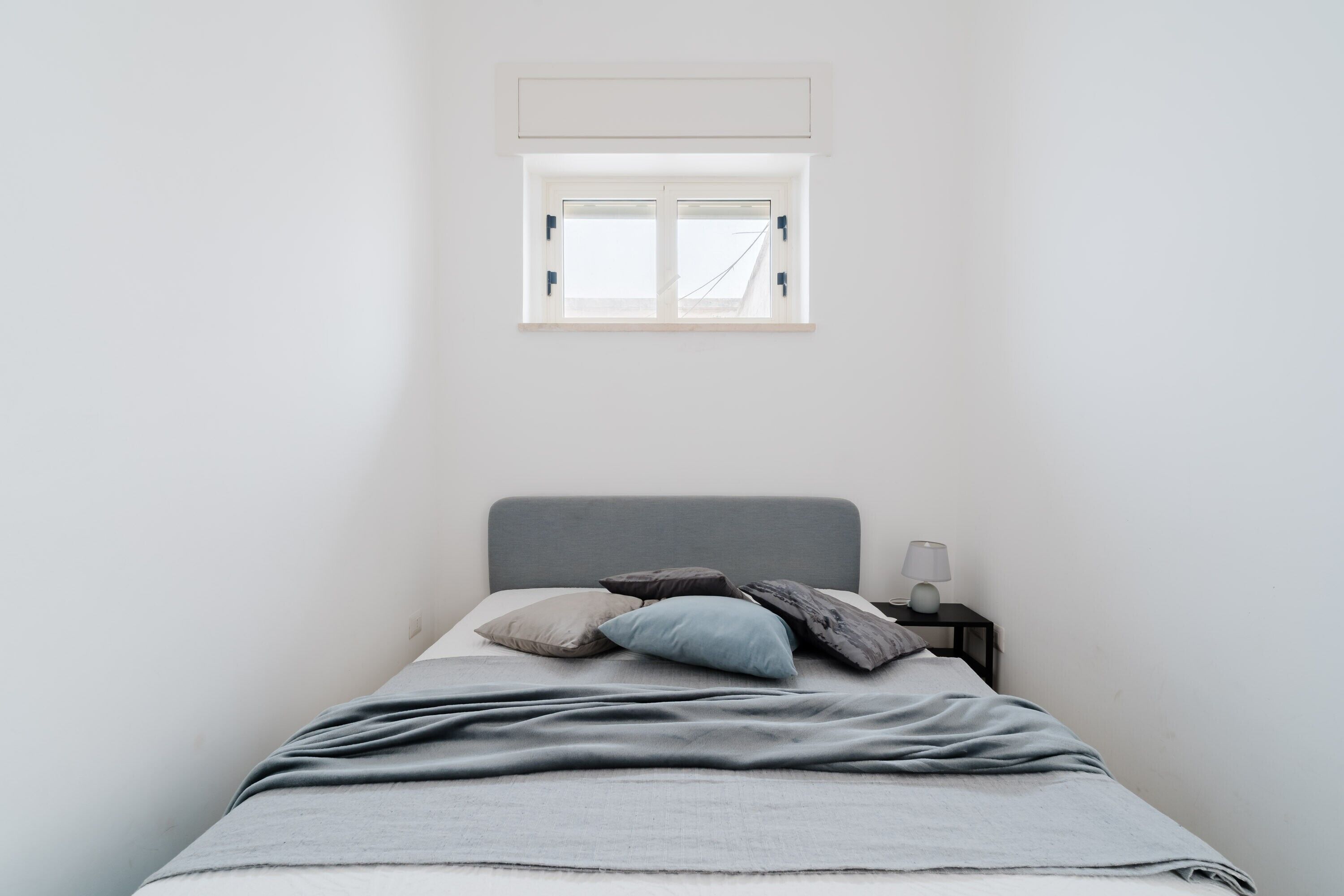 Un dormitorio con cama, almohadas, una mesita de noche, lámpara y ventana.