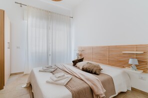 A bedroom with a bed, a bedside table with a lamp, a window with curtains, and a wooden headboard.