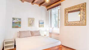 A bedroom with a wooden floor, a bed with white bedding, a nightstand, a window with a view, and three framed artworks on the wall. - Holiday Home 'Priest's Traditional Mansion' with Sea View, Shared Terrace and Wi-Fi (Corfu)
