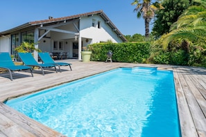 A swimming pool with lounge chairs, a covered patio, and a white building with a tiled roof. - Villa 'Charming Landes House with Private Pool, Wi-Fi and Air Conditioning' (Labenne)