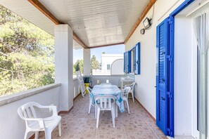 Een overdekt balkon met een tafel en stoelen, een blauwe deur en uitzicht op groen.