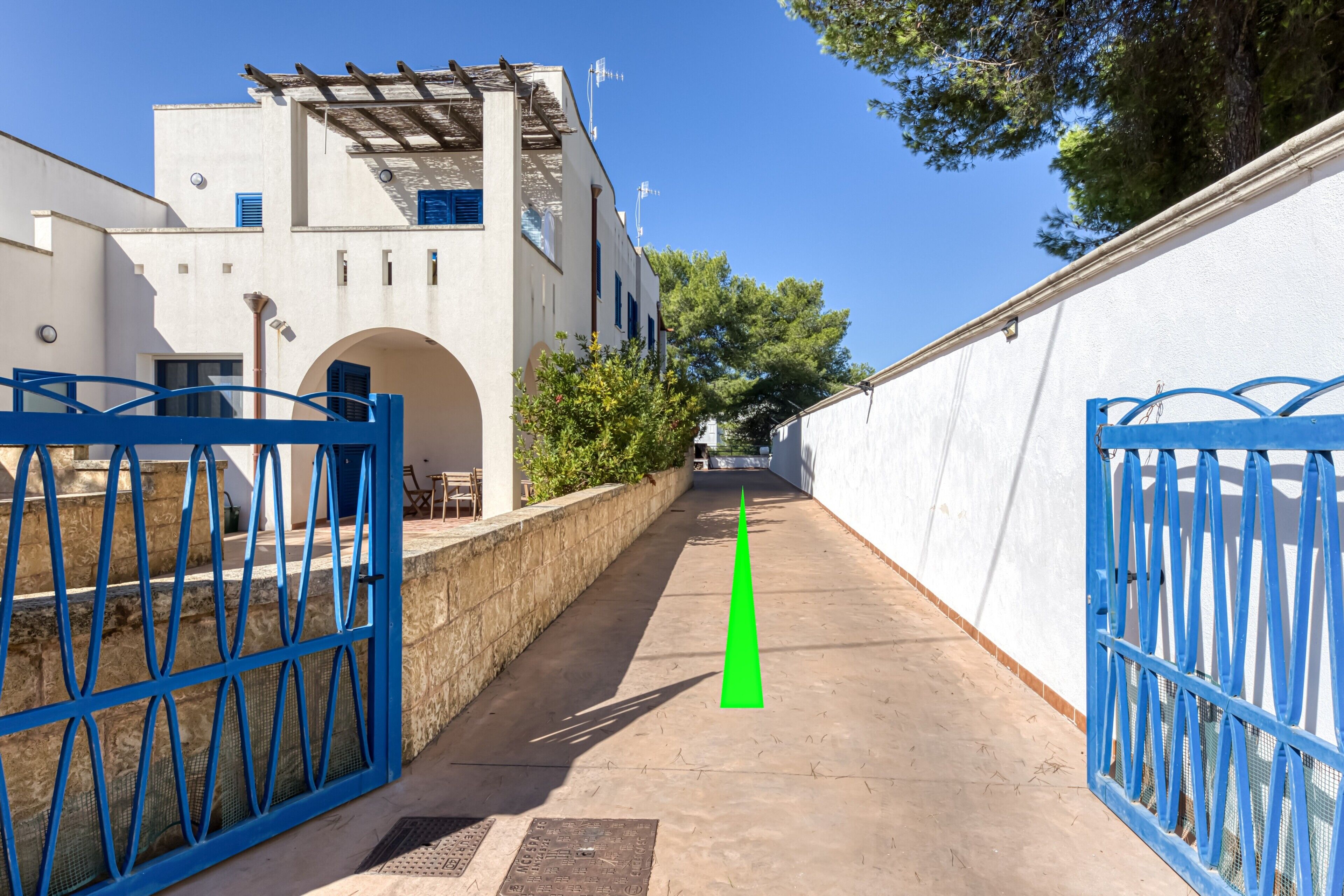 Une ruelle étroite avec un portail bleu, un bâtiment blanc doté d’un mur en pierre et un ciel d’un bleu limpide.