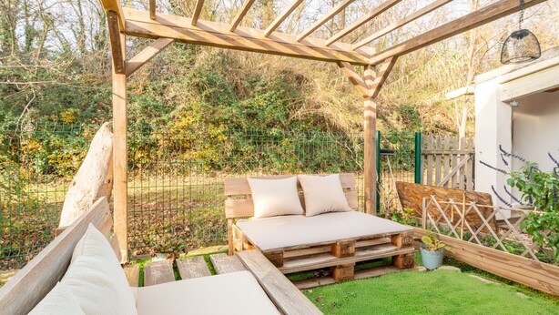 Een houten pergola met een bed en een tafel, omgeven door een groen gazon en een hek.