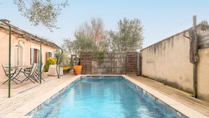 Een rechthoekig zwembad met een houten terras, een houten omheining en een gebouw met een deur en ramen.