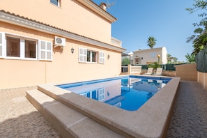 A swimming pool with a clear blue water surface, a beige building with white shutters, and a tiled deck.