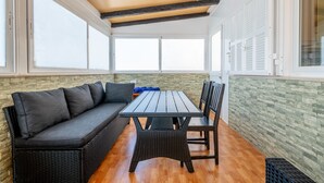 Een kamer met een donkere rieten bank, een houten tafel en stoelen, grote ramen en een stenen muur.