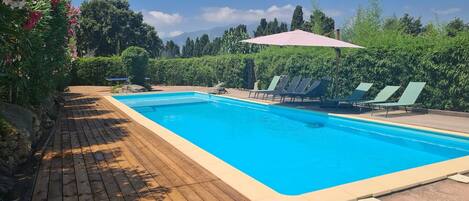 A rectangular swimming pool with a wooden deck, surrounded by greenery and a mountain backdrop.