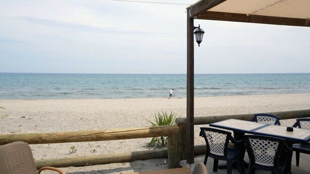 A beachside restaurant with outdoor seating, a view of the sea, and a person walking on the sand. - Villa 'Via Ferrata' with Mountain View, Private Pool and Private Terrace (Canale-di-Verde)