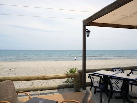 A beachside restaurant with outdoor seating, a view of the sea, and a person walking on the sand.
