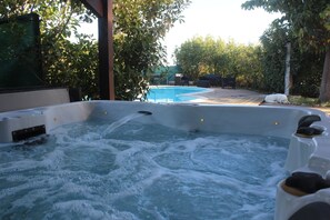 Un bain à remous avec vue sur une piscine, entouré d’arbres et doté d’un patio couvert.