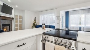 A modern kitchen with a built-in stove, white cabinetry, a flat-screen TV mounted above the fireplace, and a Christmas tree.