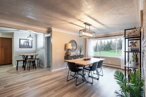 A spacious dining area with a wooden table and chairs, a hanging light fixture, and a view of the outdoors through large windows.