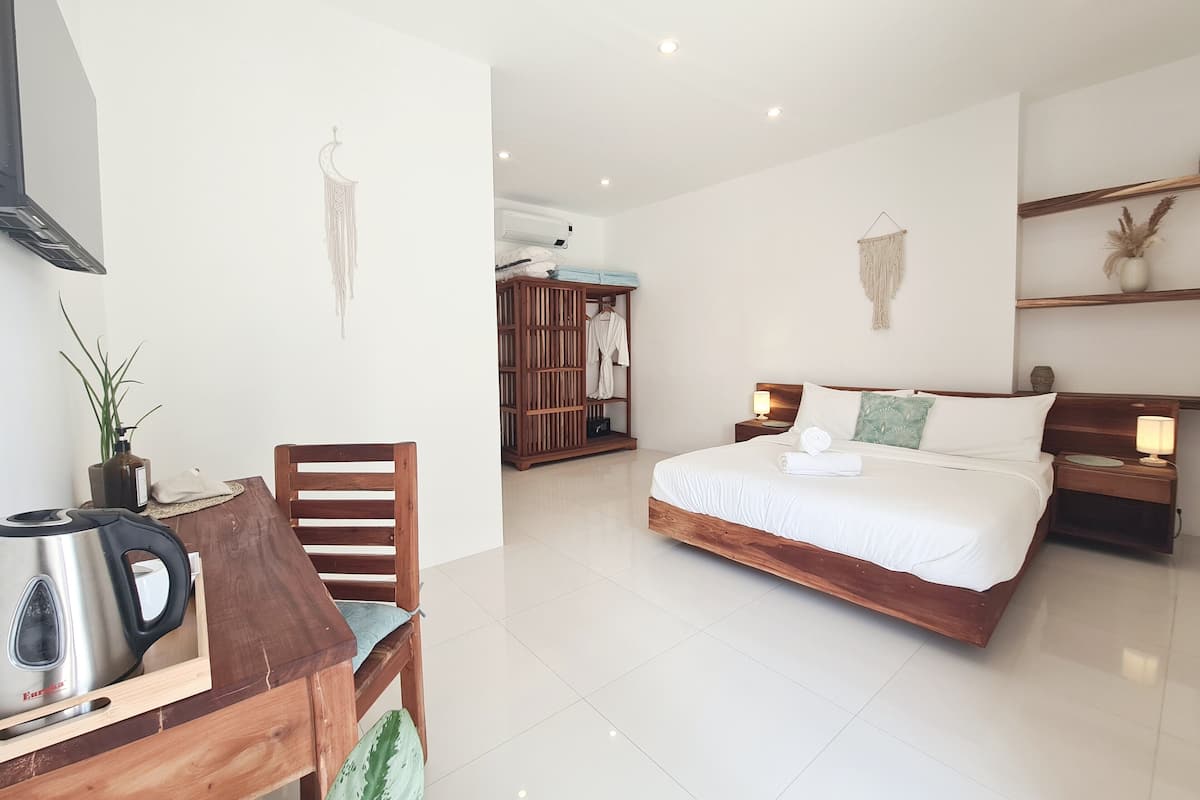 Una habitación de hotel moderna con una cama y una silla de madera, una mesita con un hervidor, un armario y un estante fijado a la pared.