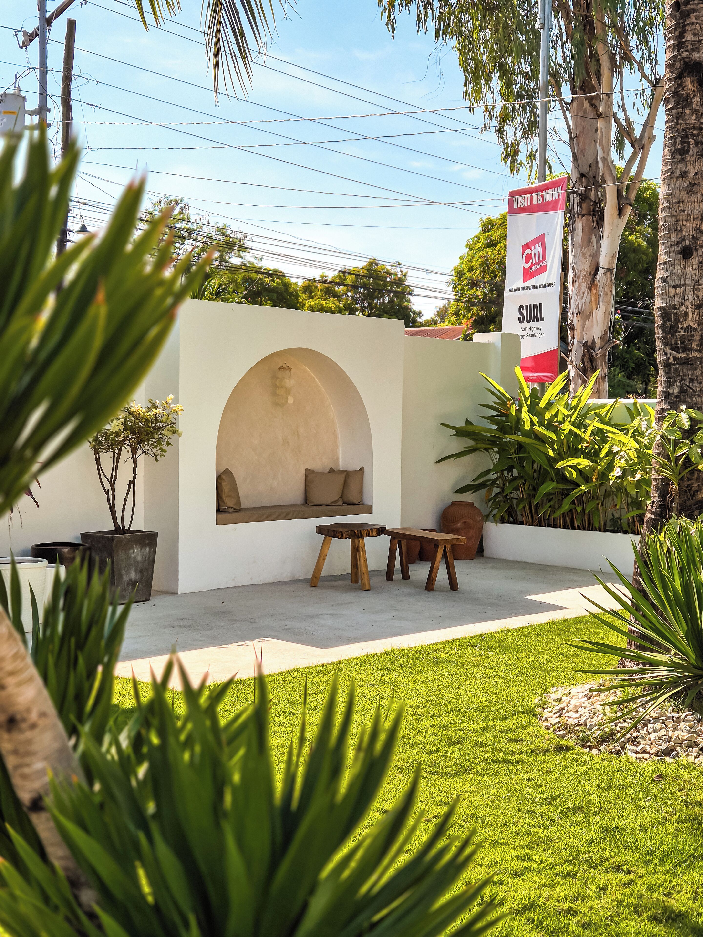 Un espace extérieur moderne, aménagé avec un banc et une table, entouré d’une végétation luxuriante et bordé d’un mur blanc percé d’une fenêtre en arc de cercle.