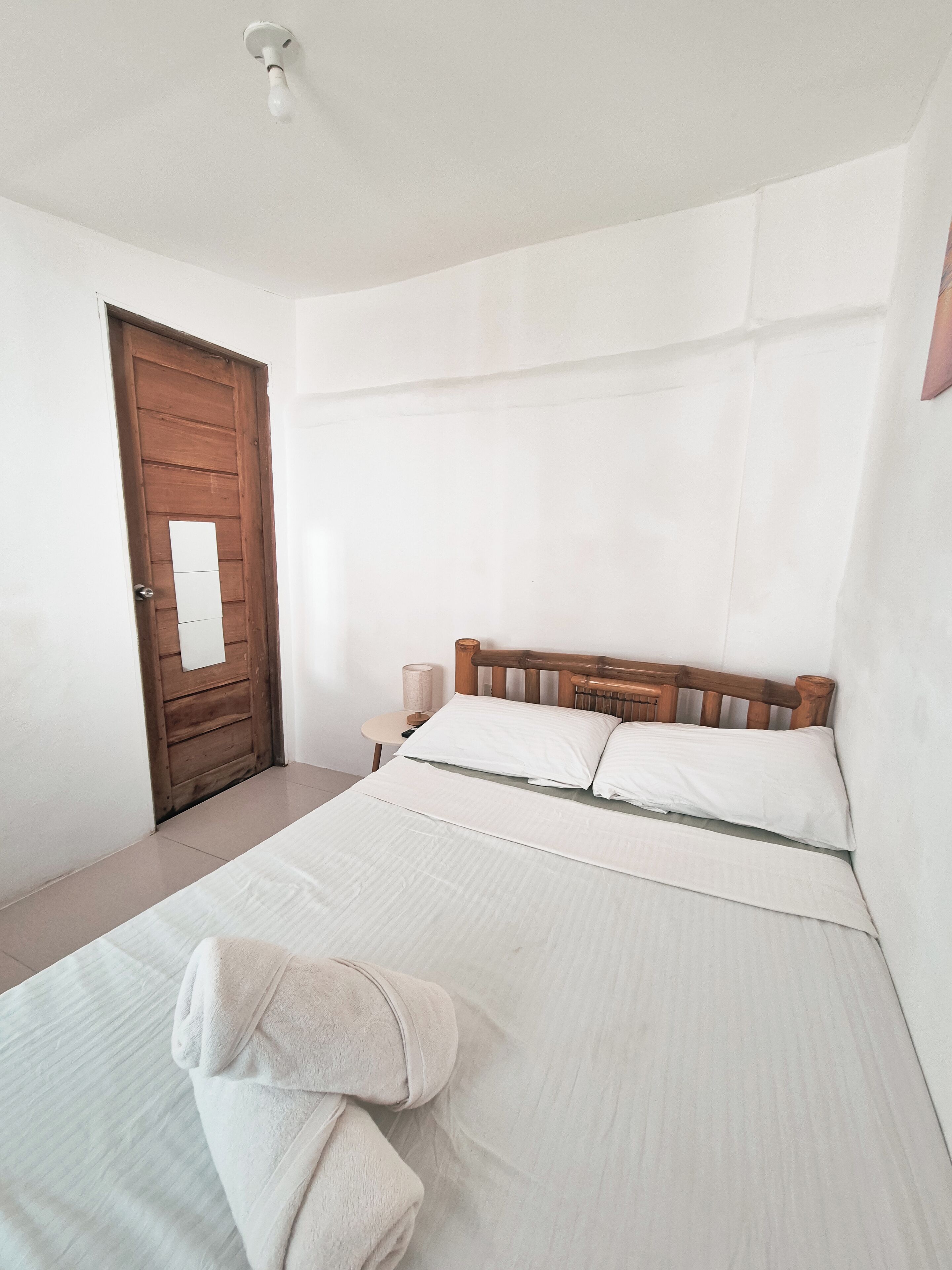 A bedroom with a wooden door, a small bedside table with a lamp, a bed with white bedding, and a rolled-up towel on the bed.