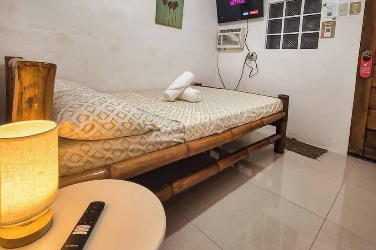 Una habitación de hotel con cama, televisor montado en la pared, una mesita con control remoto y una lámpara.