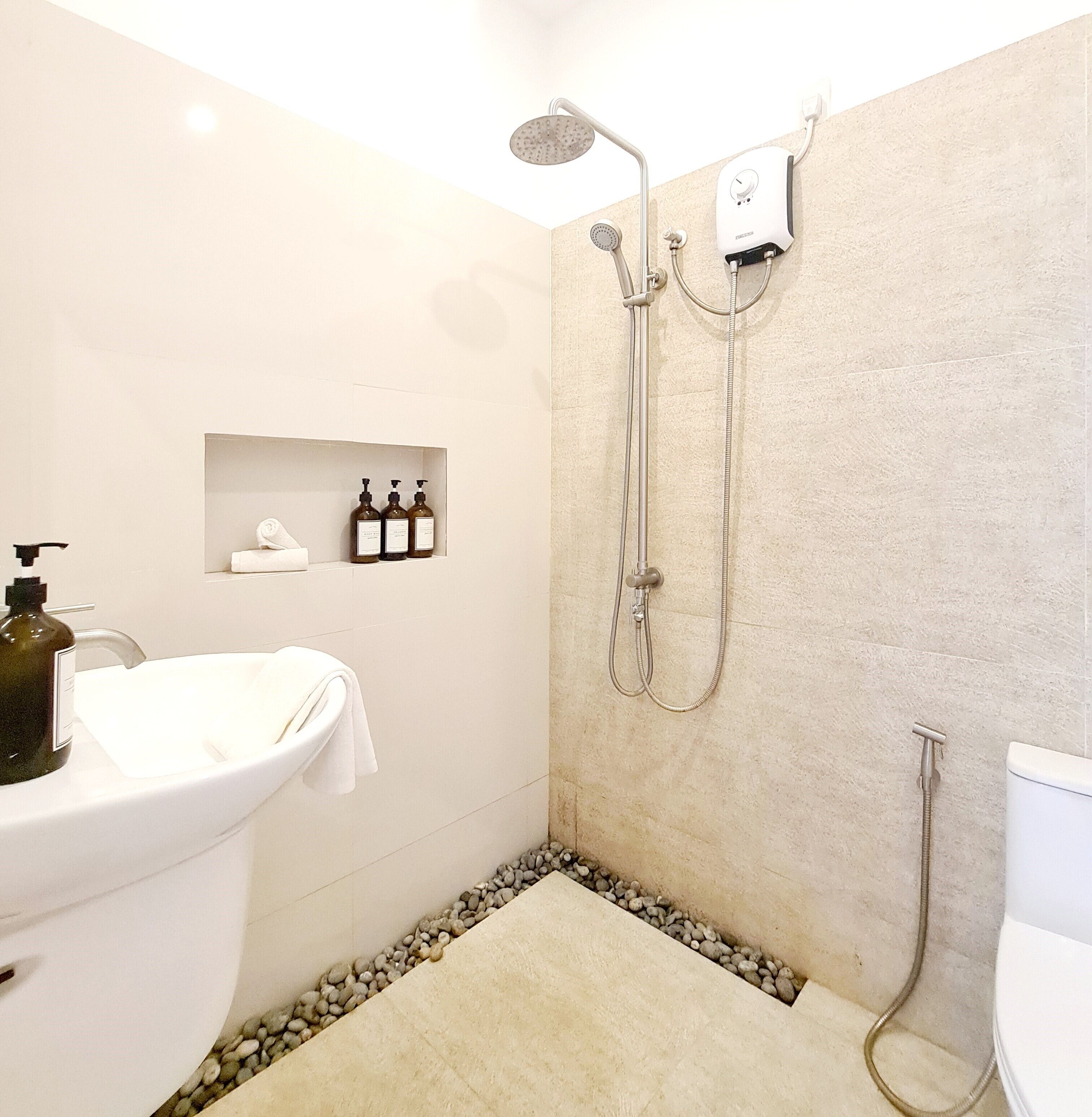 Un baño moderno con un lavamanos blanco, una ducha con cabezal de ducha manual, y un cabezal de ducha manual montado en la pared. Hay tres botellas de champú y acondicionador en un estante sobre el lavamanos.