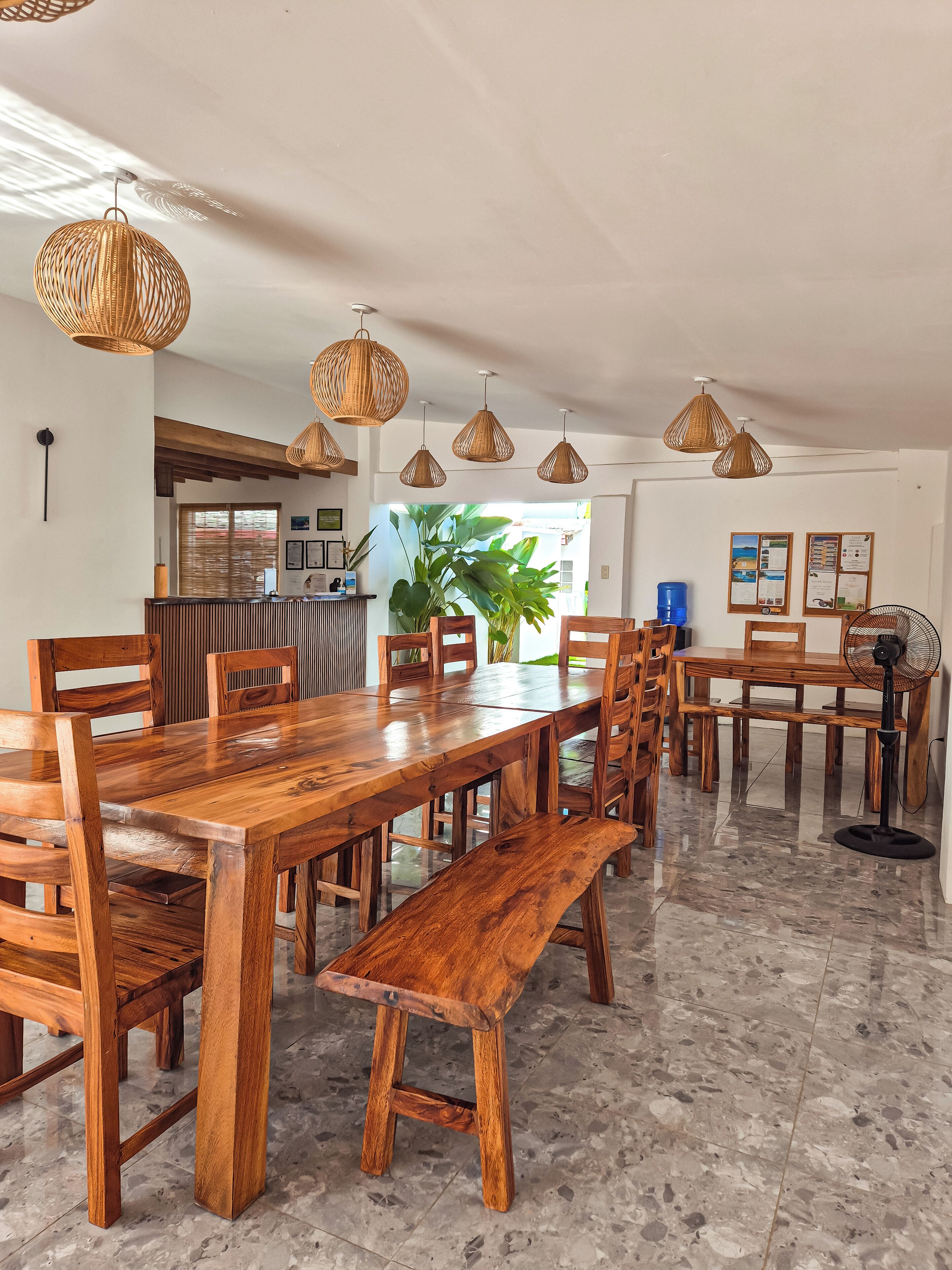 A spacious dining area with wooden tables and chairs, woven pendant lights, and a large potted plant.