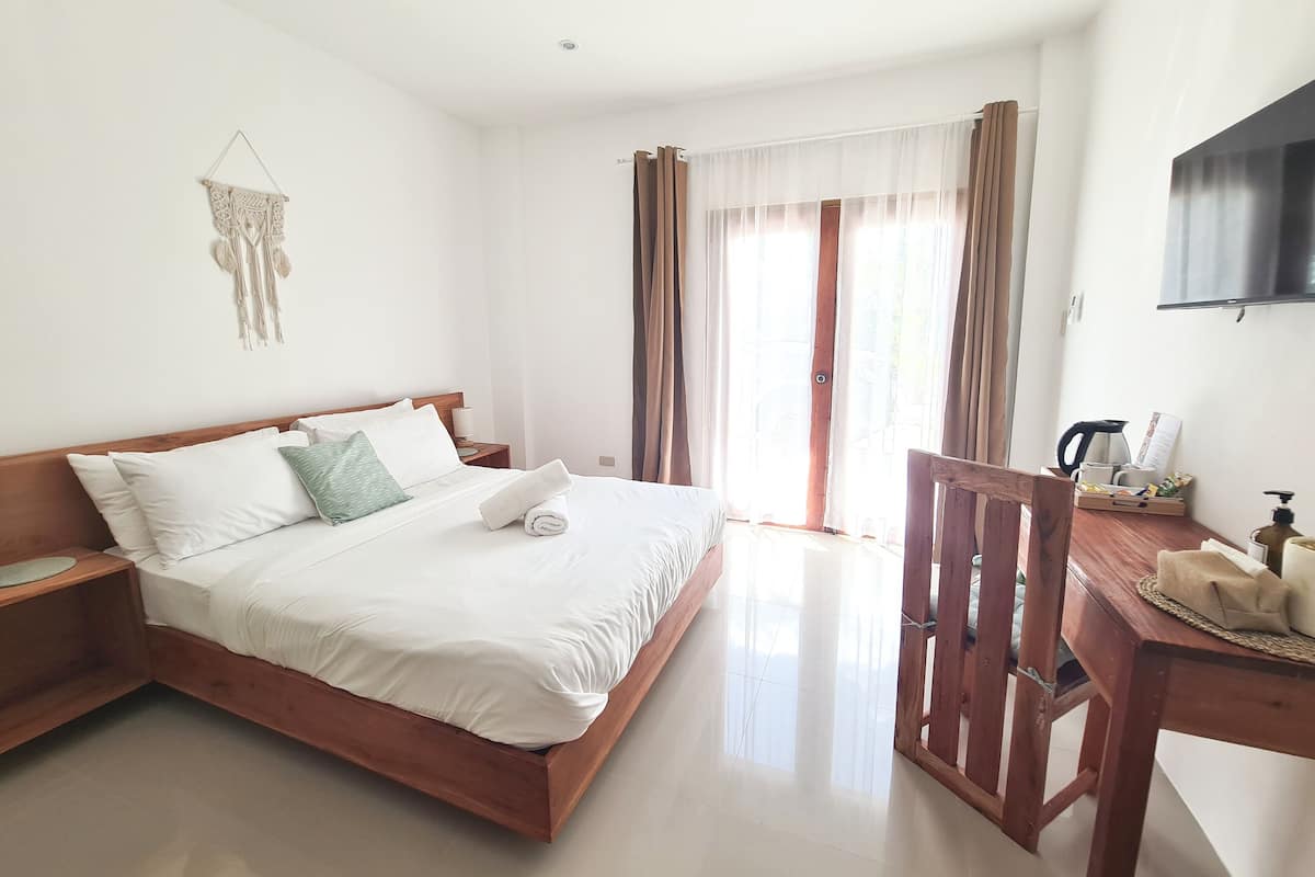 Una cama bien hecha con ropa blanca y una toalla enrollada, una mesita de noche de madera, una silla de madera, un escritorio con una cafetera y un televisor en la pared.