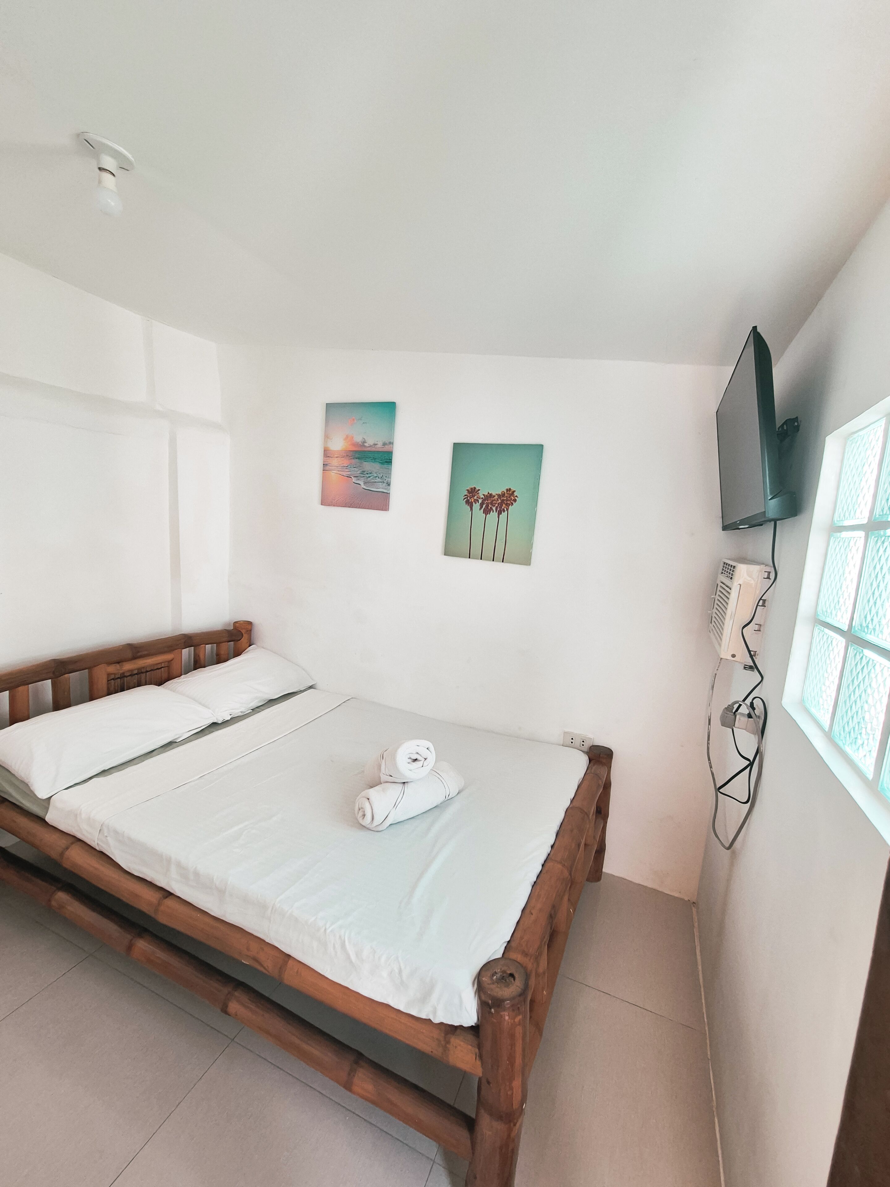 Ein Einzelbett mit Holzrahmen und weißer Bettwäsche, ein an der Wand befestigtes Fernsehgerät und zwei kleine Wandbilder mit Sonnenuntergang und Palmen.