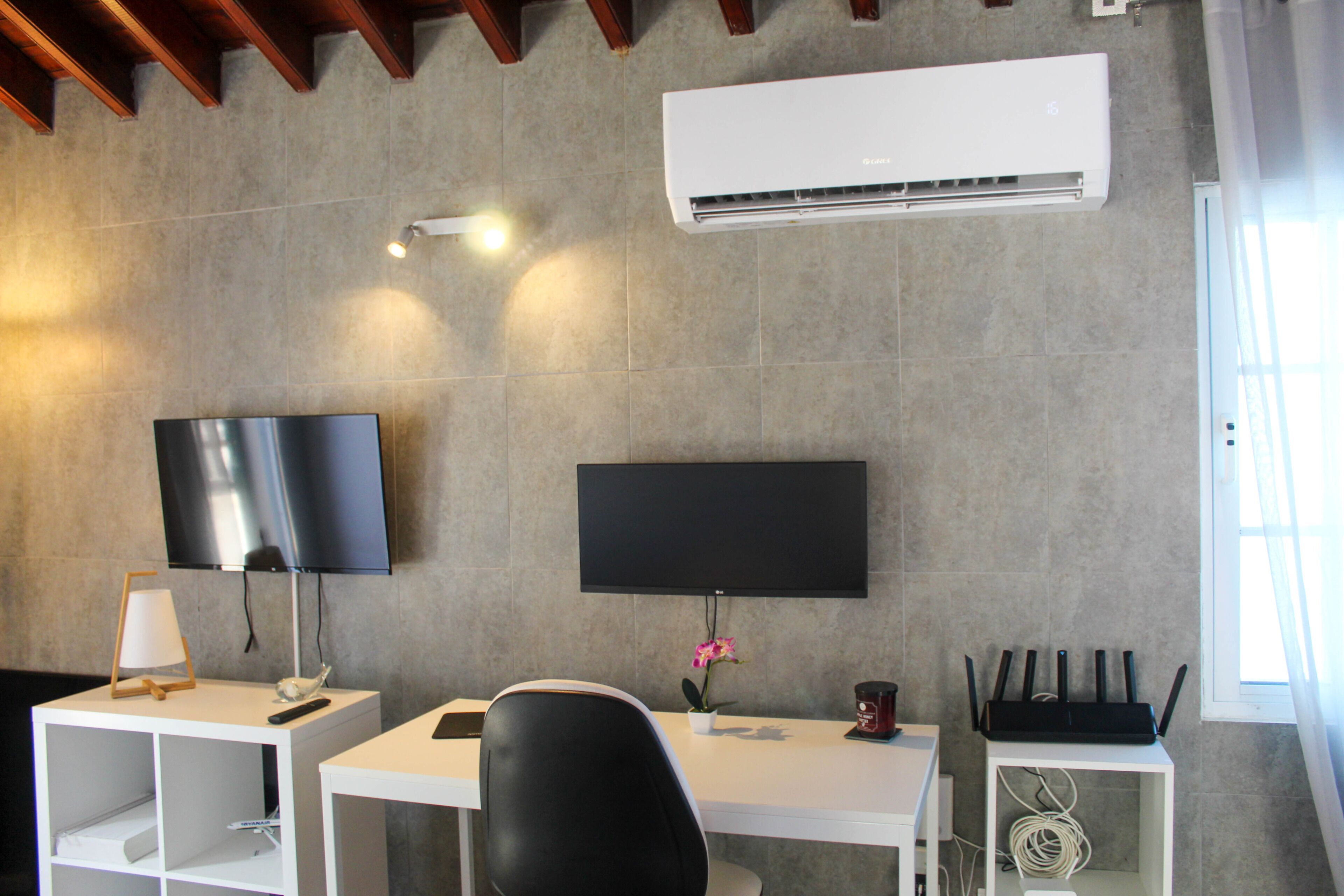 Habitació moderna amb televisió de pantalla plana, escriptori, cadira, taula petita amb làmpada i aire condicionat a la paret.