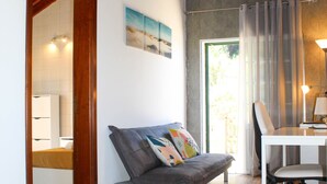 A modern living room with a wooden floor, a grey sofa with colorful cushions, a white table with a chair, and a large window with curtains. - House 37North (Ponta Delgada)