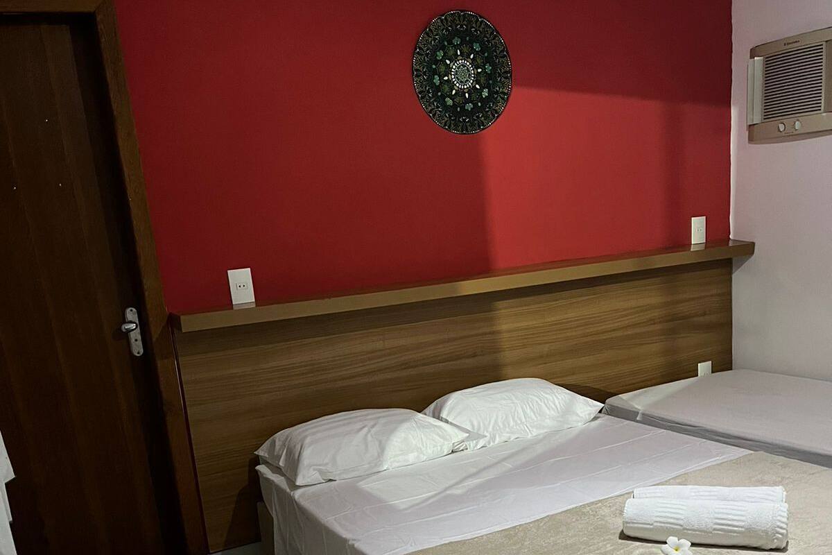 A hotel room with a wooden headboard, two beds, a red accent wall, a decorative plate, and a wall-mounted air conditioner.