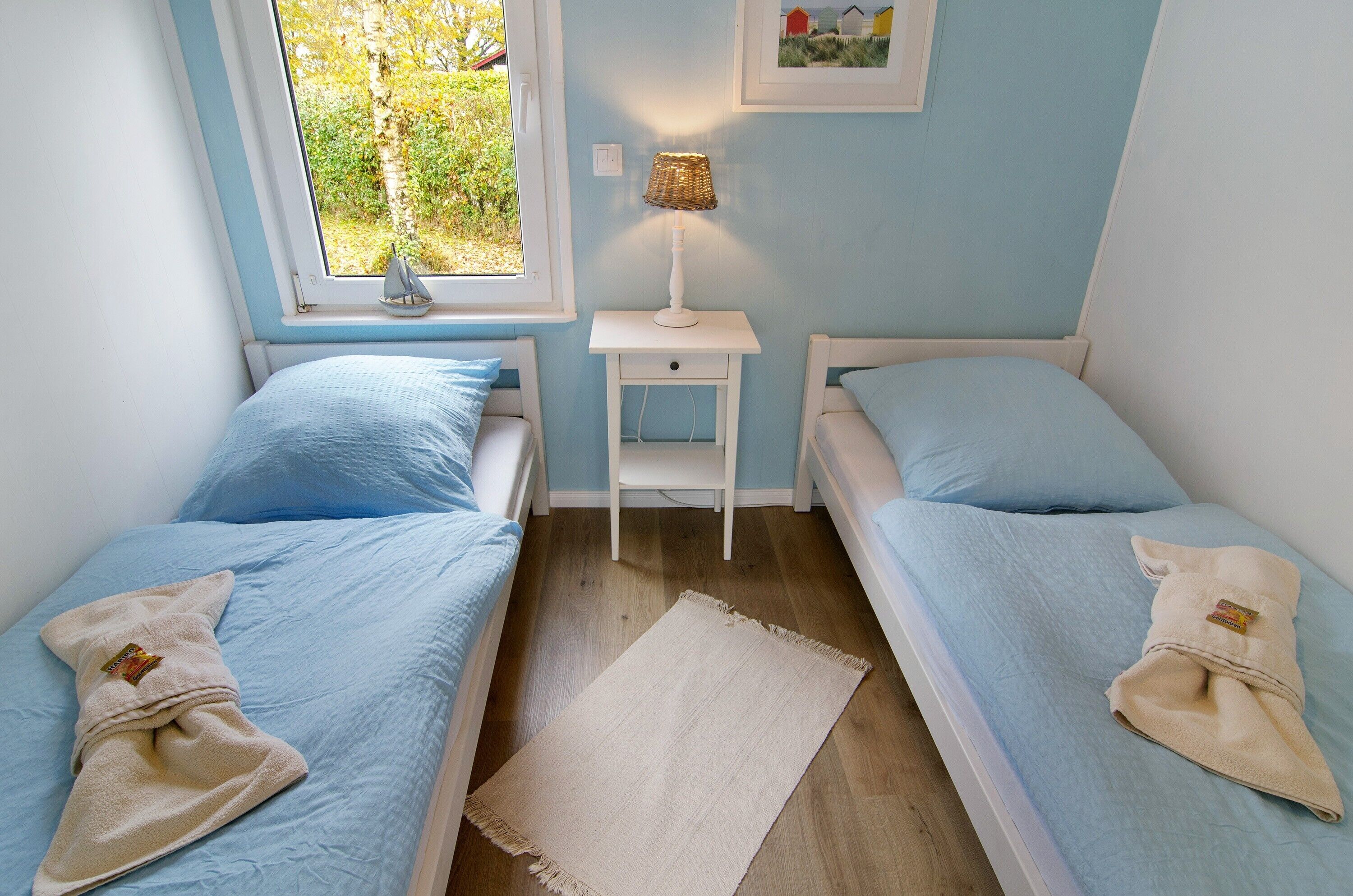 A bedroom with two beds, a nightstand, a lamp, and a window with a view of greenery.