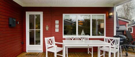 A red wooden house with a white door, a white table with chairs, and a large window.