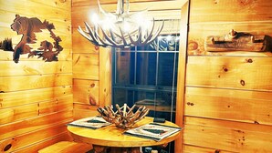 A wooden cabin interior with a round wooden table, two stools, a chandelier, and a window with a view of trees. - Cozy Bear: A Dreamy Cabin Hideout for Two, Gatlinburg & Pigeon Forge - SPECIALS (Pigeon Forge)