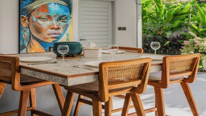Un comedor con una mesa dispuesta para cuatro personas, rodeada de sillas de madera con respaldo trenzado. Hay tres lámparas colgantes sobre la mesa, y un cuadro grande cuelga en la pared a la izquierda.