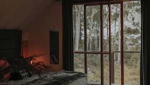 A bedroom with a large bed, a wooden floor, a patterned rug, and a view of trees through a large window.