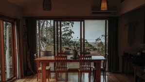 A dining area with a wooden table and chairs, large windows with curtains, and a view of trees and a distant landscape.