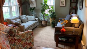 A living room with a sofa, a floral armchair, a coffee table, and a lamp. There are potted plants and framed pictures on the walls.
