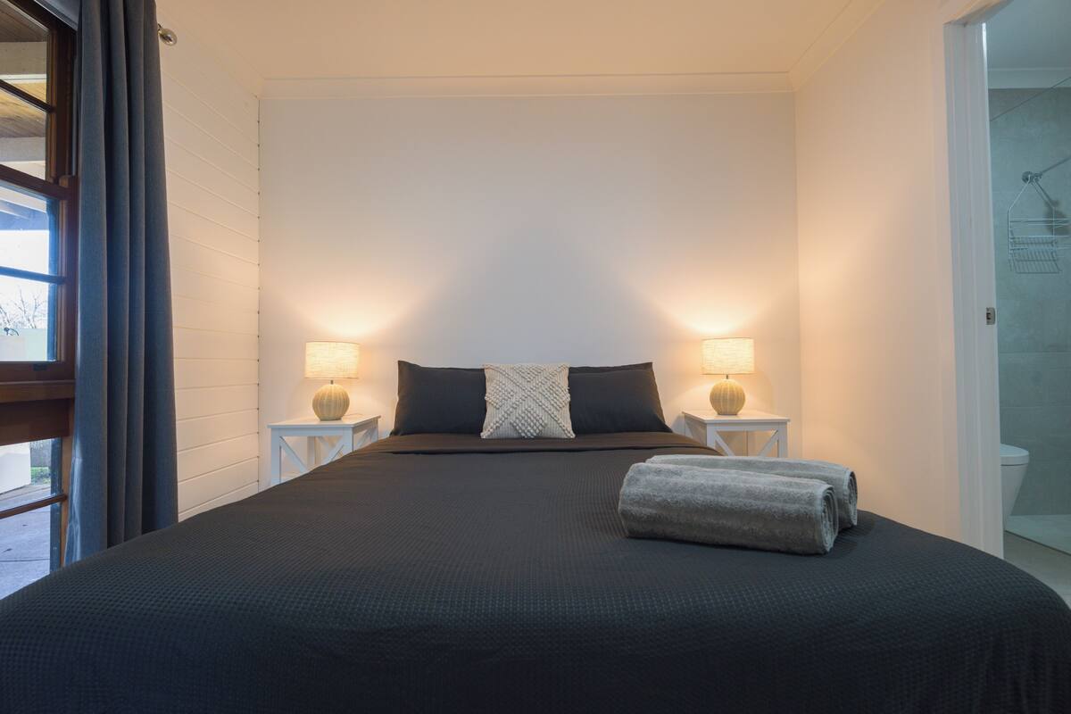 Una cama bien hecha con un edredón oscuro y dos almohadas, con dos lámparas de noche a cada lado sobre mesitas blancas.
