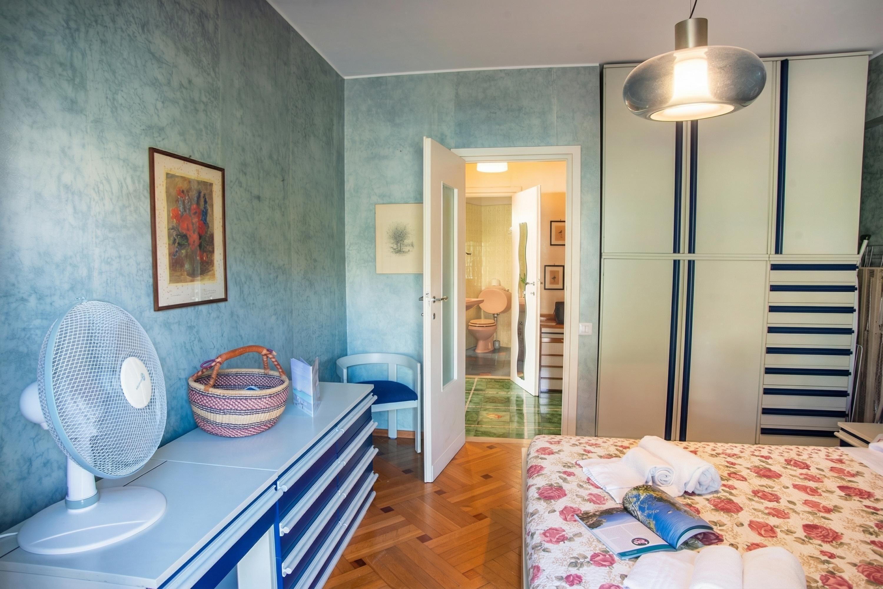 A bedroom with a bed, a desk with a fan, a chair, a wardrobe, and a bathroom visible through an open door.