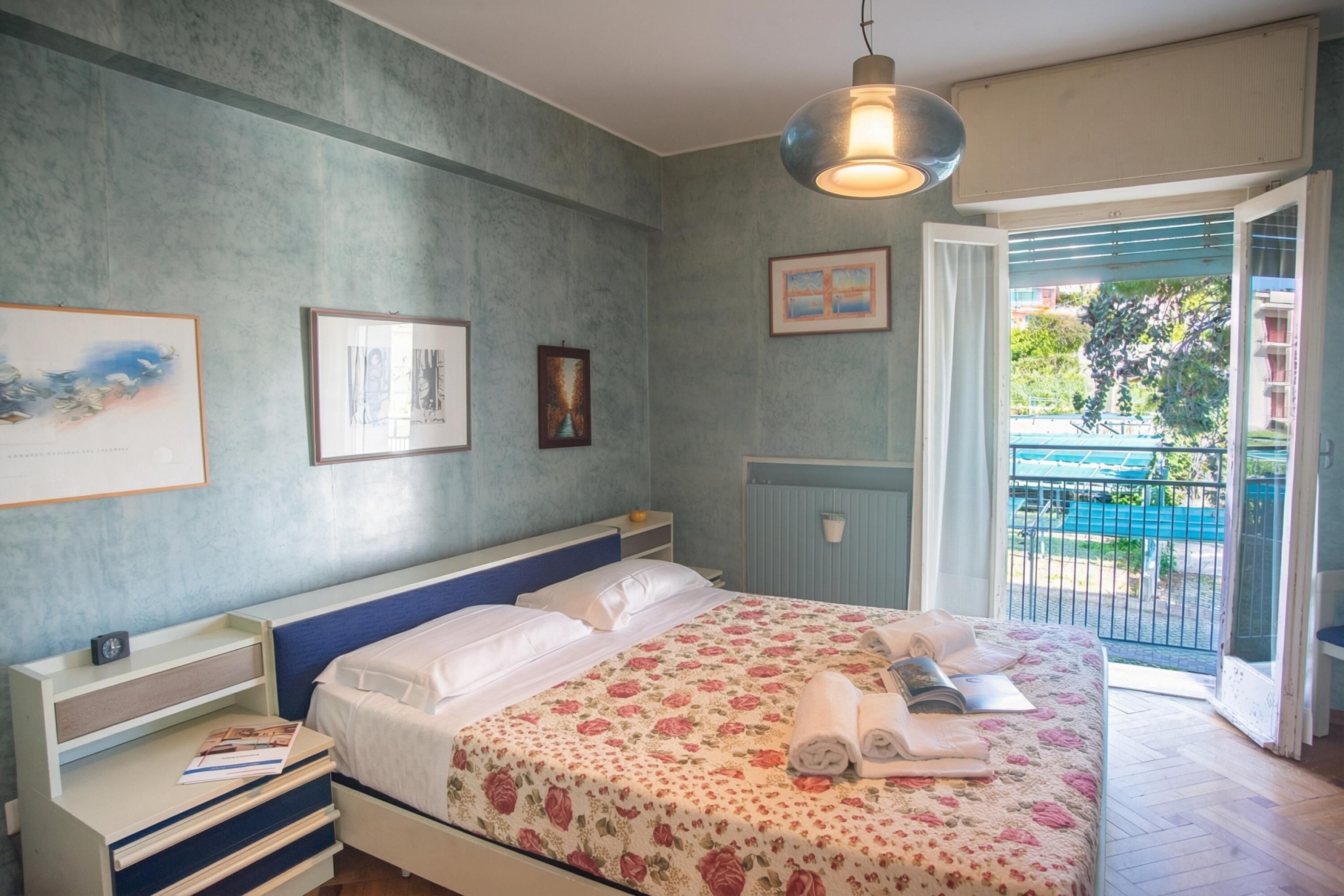 A bedroom with a bed, bedside tables, a window with a view, and a door leading to a balcony.