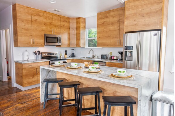 A modern kitchen with wooden cabinets, stainless steel appliances, a marble island with stools, and a view of greenery outside. - Cozy and Gorgeous 2BR 1BA Near Amazing Downtown (San Antonio)
