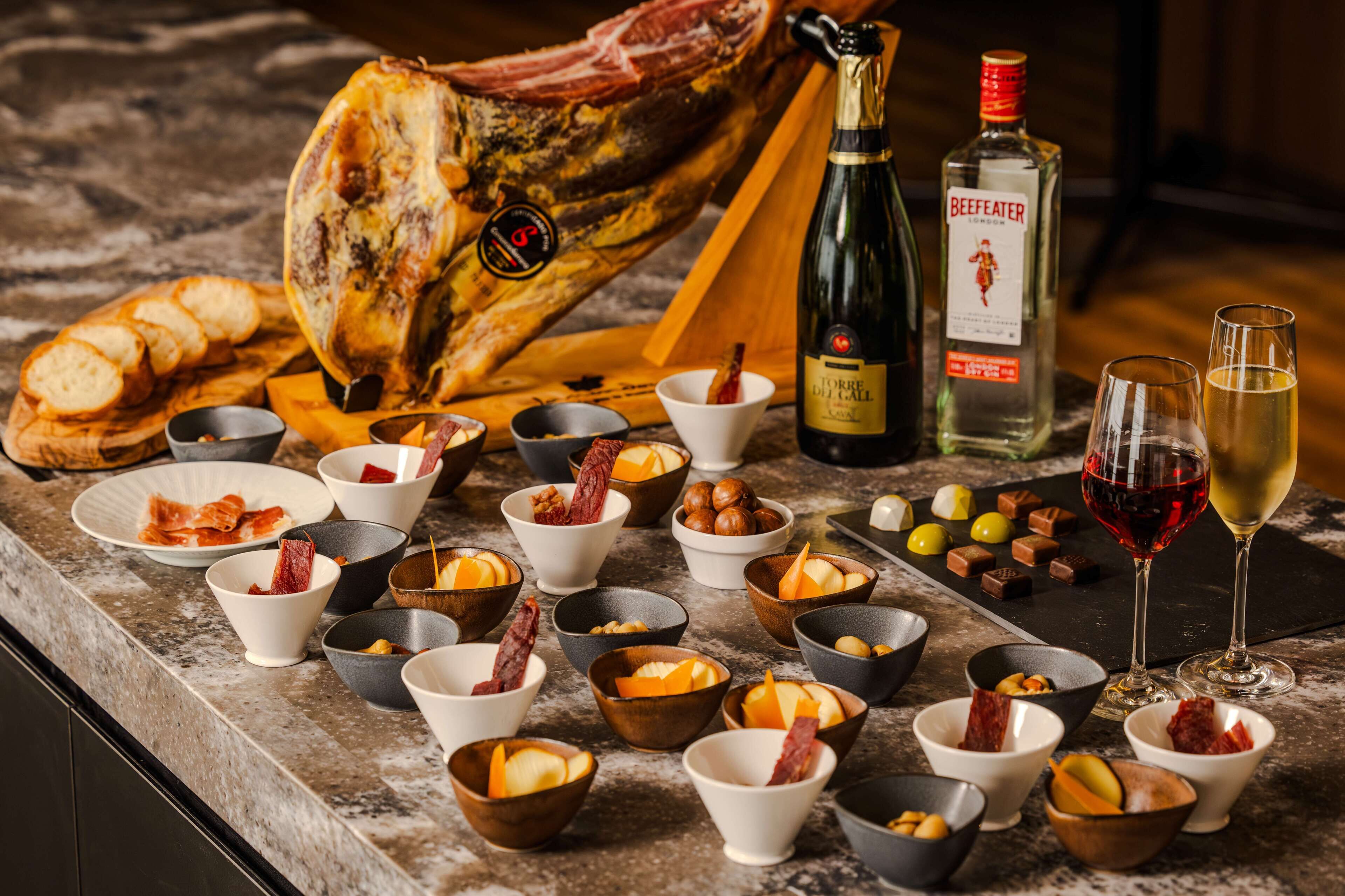 Una tabla de embutidos con diversos tipos de carne, quesos y acompañamientos, una botella de vino y copas sobre una encimera de mármol.