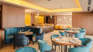 A modern dining area with blue upholstered seating and round tables, featuring a central bar and ambient lighting.