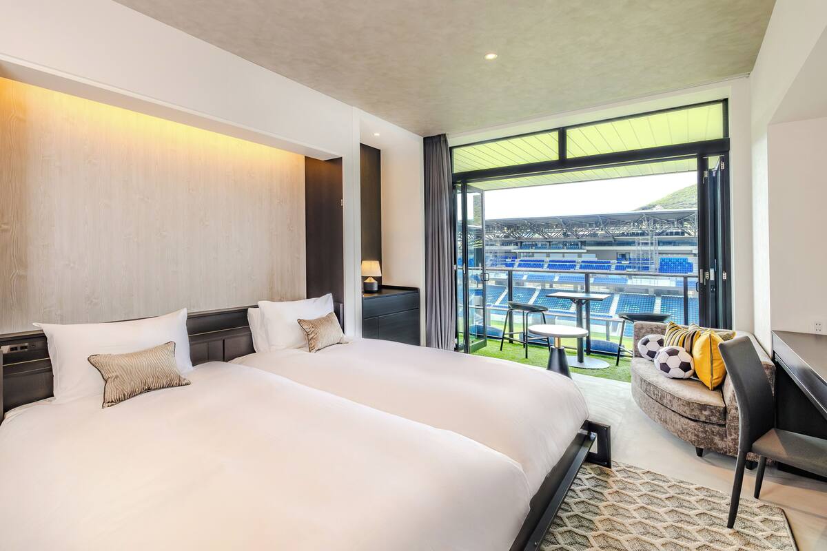 Ett modernt hotellrum med en stor säng, en sittgrupp med soffa och fåtöljer samt en balkong med utsikt över en stadion.