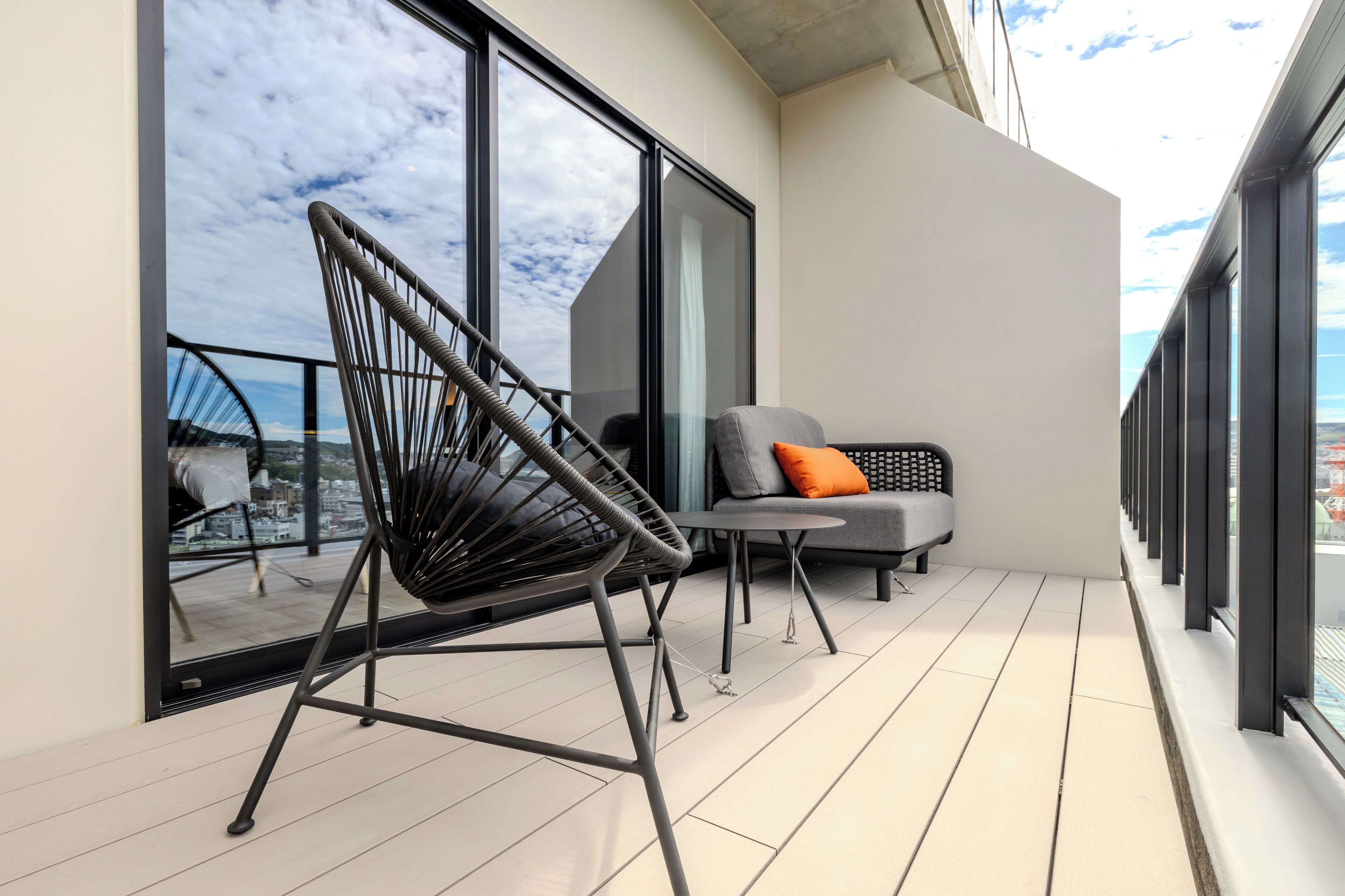 A balcony with a black chair, a grey sofa with an orange pillow, and a small table.