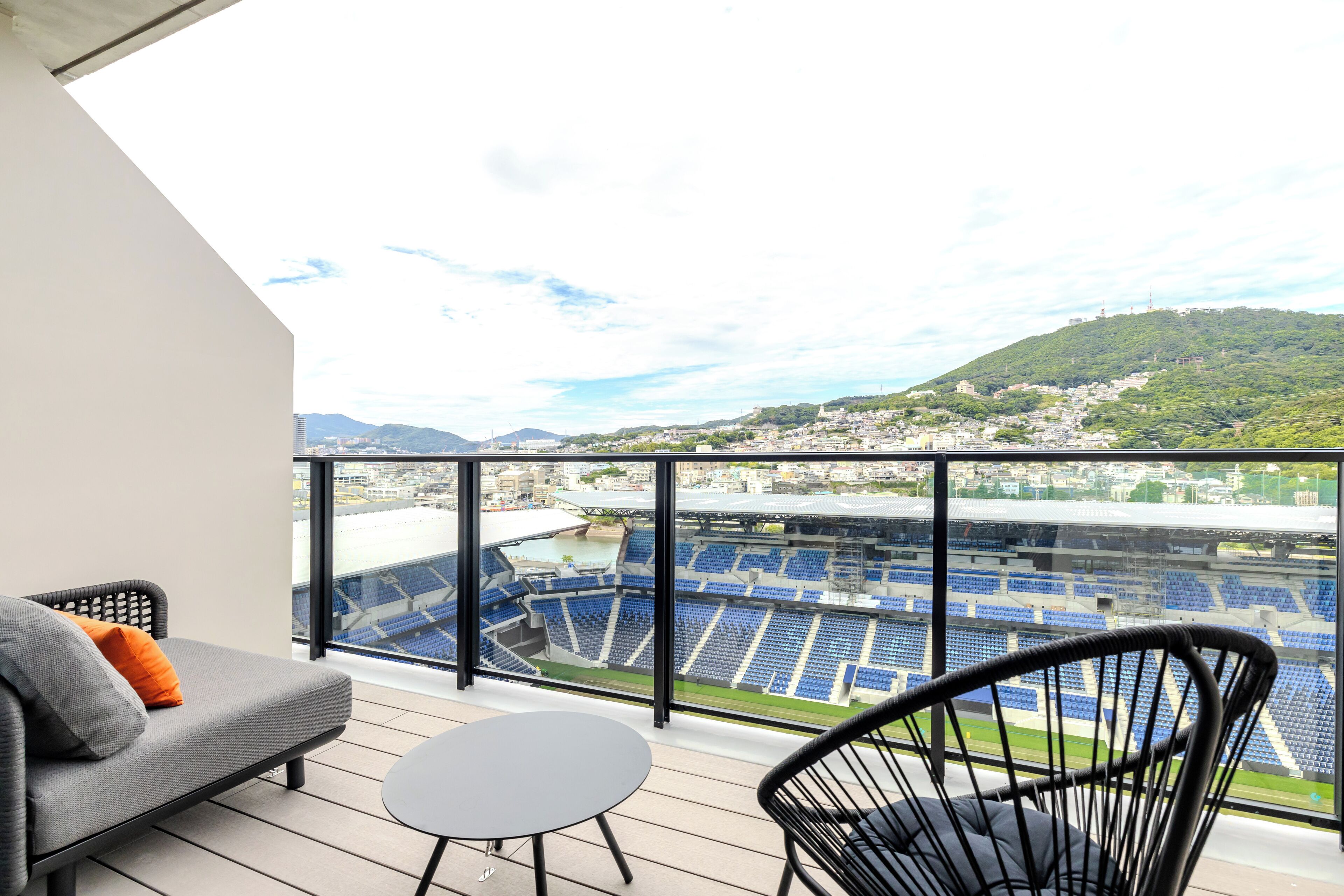 A balcony with a view of a stadium, a grey sofa, a round table, and two wicker chairs.